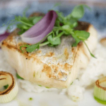 An elegantly plated dish featuring a perfectly cooked portion of hake, garnished with vibrant green herbs and vegetables on a white plate, set against a blurred background for aesthetic presentation.