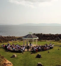 An outdoor wedding ceremony setup with white chairs, a floral arch, and an ocean view under a clear blue sky.