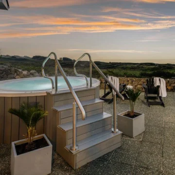 Outdoor hot tub overlooking scenic ocean view, surrounded by rocks and greenery. Adirondack chairs are placed nearby for relaxation. A serene and inviting atmosphere is depicted.