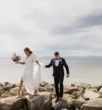 A beautifully set wedding reception area with elegant table arrangements, floral centerpieces, white chairs, and a scenic backdrop of the ocean and clear blue skies.