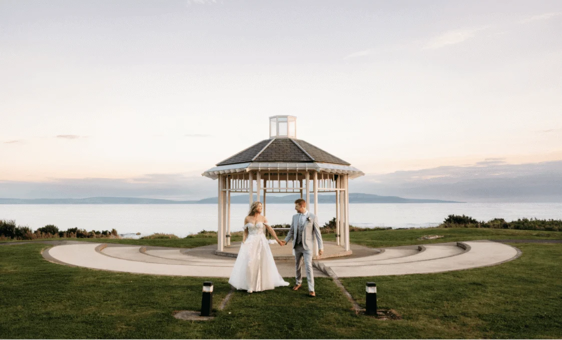 A charming wedding ceremony setup with a white gazebo, floral decorations, and scenic ocean view in the background on a sunny day.