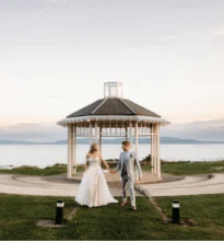 A charming wedding ceremony setup with a white gazebo, floral decorations, and scenic ocean view in the background on a sunny day.