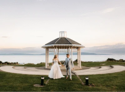 A charming wedding ceremony setup with a white gazebo, floral decorations, and scenic ocean view in the background on a sunny day.