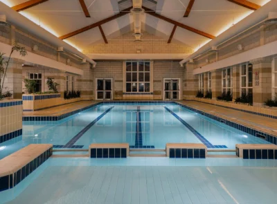 Indoor swimming pool with clear blue water, surrounded by windows and modern furnishings, set in a leisure center featuring gym equipment and potted plants for a calming atmosphere.