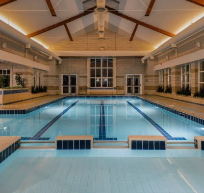 Indoor swimming pool with clear blue water, surrounded by windows and modern furnishings, set in a leisure center featuring gym equipment and potted plants for a calming atmosphere.