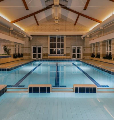 Indoor swimming pool with clear blue water, surrounded by windows and modern furnishings, set in a leisure center featuring gym equipment and potted plants for a calming atmosphere.