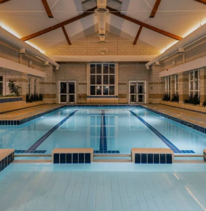 Indoor swimming pool with clear blue water, surrounded by windows and modern furnishings, set in a leisure center featuring gym equipment and potted plants for a calming atmosphere.