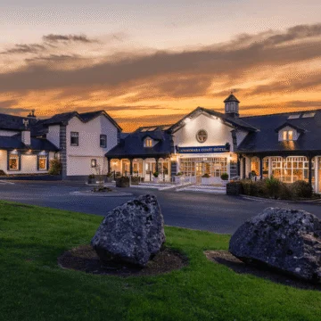 "Connemara Coast Hotel exterior, lit by a vibrant sunset. The building is elegant, surrounded by lush greenery, with a calm, reflective sea in the foreground."