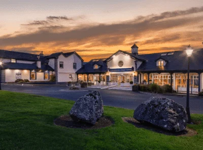 "Connemara Coast Hotel exterior, lit by a vibrant sunset. The building is elegant, surrounded by lush greenery, with a calm, reflective sea in the foreground."