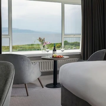 Elegant hotel room with modern furniture, large bed, and floor-to-ceiling windows showcasing a stunning ocean view and tranquil seascape; soft lighting creates a warm, inviting atmosphere.