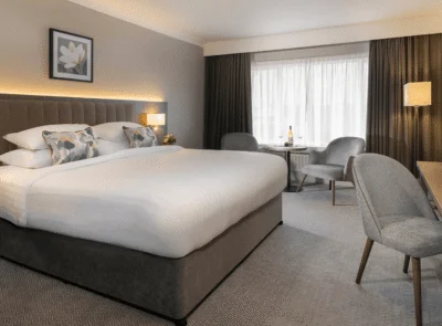 A cozy hotel room with a king-sized bed, modern furnishings, artwork on walls, and a view of the ocean through a large window.