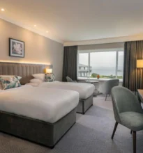 Two twin beds with white linens in a hotel room, large window displaying a scenic ocean view. Warm-toned curtains and bedside lamps create a calming, welcoming atmosphere.