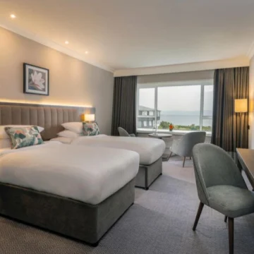 Two twin beds with white linens in a hotel room, large window displaying a scenic ocean view. Warm-toned curtains and bedside lamps create a calming, welcoming atmosphere.