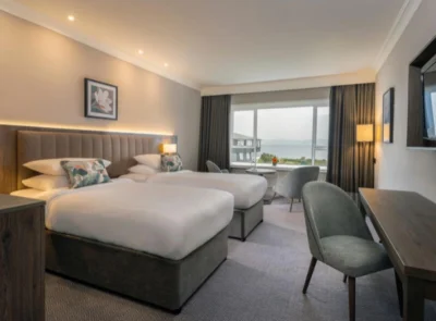 Two twin beds with white linens in a hotel room, large window displaying a scenic ocean view. Warm-toned curtains and bedside lamps create a calming, welcoming atmosphere.