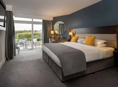 A cozy hotel room with a double bed, green accent wall, large window, wooden furnishings, and floral decor, leading to a garden patio.